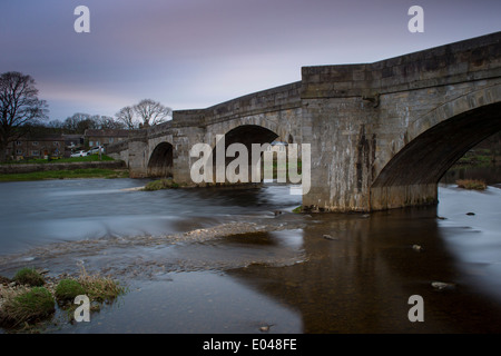 Blick auf die Steinerne Brücke über dem Fluss Wharfe durch Burnsall Dorf an einem Frühlingsabend unter blau rosa Himmel fließend - Yorkshire Dales, England, UK. Stockfoto