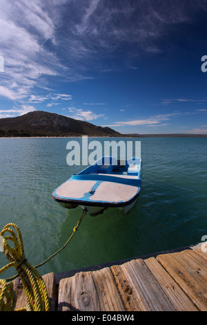 Ein kleines Ruderboot festgemacht an einem Steg an einer Lagune. Das Seil ist gelb, der Himmel blau. Stockfoto
