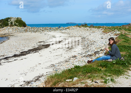 Frau sitzt SMS auf Handy am Strand von St. Agnes, Cornwall, Scillies, Isles of Scilly im April Stockfoto