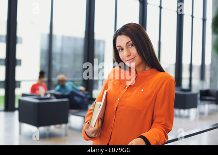 Attraktive junge Studentin mit Büchern unter dem Arm in der Schule. Kaukasische junge Frau auf dem Campus Flur mit Menschen. Stockfoto