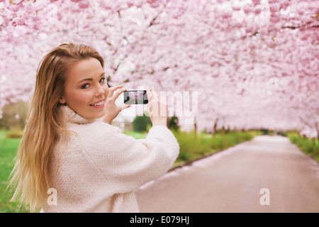 Junge Frau mit ihrem Smartphone, um Bilder von den Pfad und Kirsche Blüten-Baum im Park, junge, weibliche Blick in die Kamera zu erfassen. Stockfoto