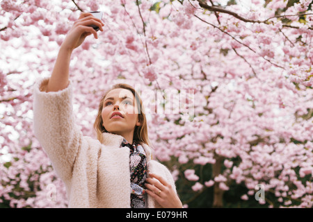 Schöne junge Dame, die Natur mit ihrem Handy zu fotografieren. Hübsche junge Frau im Frühjahr blühen Park fotografieren. Stockfoto