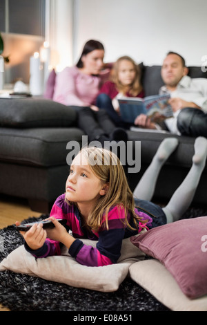 Mädchen vor dem Fernseher auf Boden mit Familie im Hintergrund Stockfoto