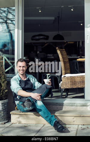 Porträt von zuversichtlich Besitzer mit Kaffeetasse sitzen am Eingang des Cafés Stockfoto