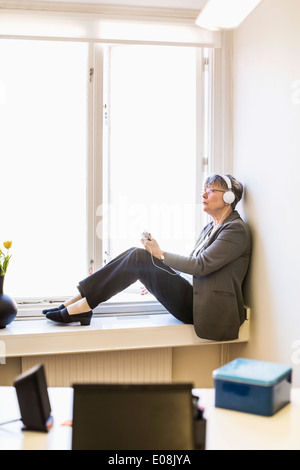 Gesamte Länge der Geschäftsfrau Musik hören beim Sitzen auf der Fensterbank Stockfoto