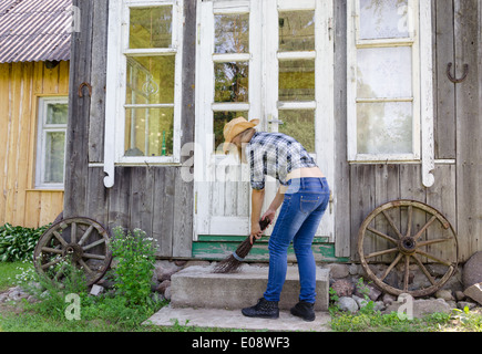 Arbeiter Frau in Jeans Hemd und Hut Tabula Rasa Treppen mit hölzernen Besen in ländlichen Hof Hof. Stockfoto