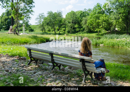 schöne Frau, die auf weißen Bank neben einem schnell fließenden Bach sitzen sieht bewundern rund Natur Stockfoto