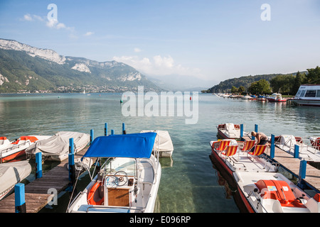 Panoramablick auf den See von Annecy, Frankreich, mit verankerten Booten und Tretbooten an einem Seeufer, Bergen und blauem Himmel im Hintergrund an einem sonnigen Tag Stockfoto