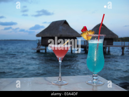 Flitterwochen. Zwei Cocktails auf einem Tisch in der Abenddämmerung mit im Hintergrund strohgedeckten Wasserbungalows auf einer tropischen Insel. Stockfoto