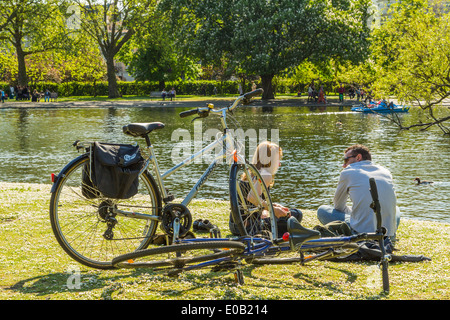 Ein junges Paar mit Fahrrädern sitzt auf dem Rasen, entspannt und unterhält sich an einem sonnigen Sommertag am See im Regents Park, London, Großbritannien Stockfoto