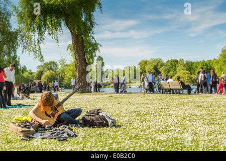 Eine junge Frau sitzt und Gitarre spielen neben dem See im Regents Park London England Großbritannien Stockfoto