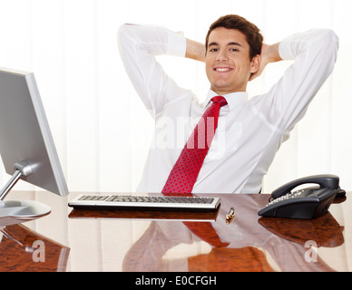 Ein erfolgreicher junger Manager sitzt mit Schreibtisch und lächelt., Ein Erfolgreicher Junger Manager Sitzt Bei Schreibtisch Und Laechelt. Stockfoto