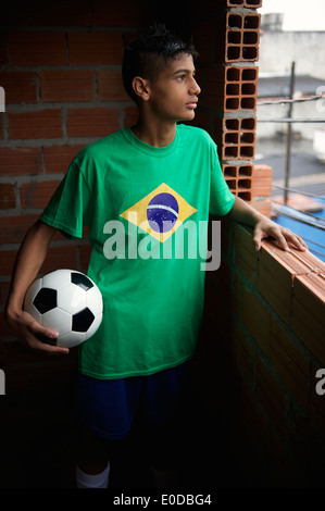 Profilbildnis ernst brasilianischen jungen Mannes trägt Brasilien Flagge T-shirt Stand halten Fußball schaut aus dem Fenster Stockfoto