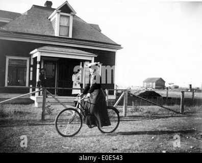Eine Frau, die in der 28. Straße Fahrrad fährt, auf einem historischen Foto festgehalten. Das Bild zeigt ihre gemütliche Fahrt und die Umgebung im frühen 20. Jahrhundert, einschließlich Straßen, Gebäuden und einer familiären Umgebung im Hintergrund. Stockfoto