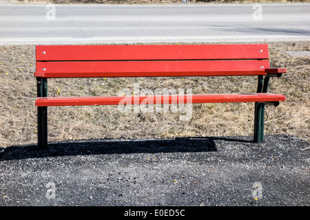 An einem sonnigen Tag lädt eine pulsierende rote Parkbank neben einer ruhigen Straße zum Entspannen ein. Stockfoto