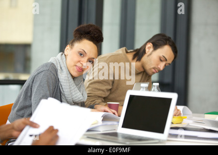 Selbstbewusste junge Frau sitzt am Tisch, Blick in die Kamera. Studierende mit Büchern und Laptop in der Bibliothek. Stockfoto