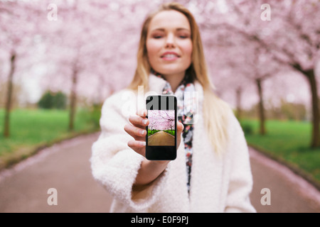 Junge Frau zeigt ein Foto auf ihrem Smartphone an Sie. Weibchen im Frühlingspark Bilder auf ihrem Mobiltelefon anzeigen. Stockfoto