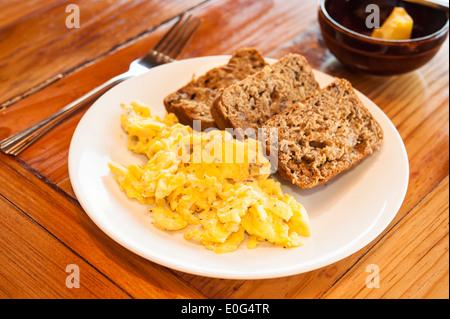 Teller mit Rührei und Bananenbrot auf einem Holztisch mit einer Schüssel mit butter Stockfoto