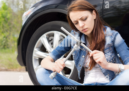 Unwissende Frau Stirnrunzeln bei einem Rad-Schraubenschlüssel, als sie auf der Straße sitzt neben ihr Auto nach einem Zusammenbruch auf Hilfe warten. Stockfoto