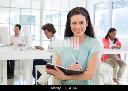 Attraktive Geschäftsfrauen in Notebook mit Kollegen hinter ihr schreiben Stockfoto