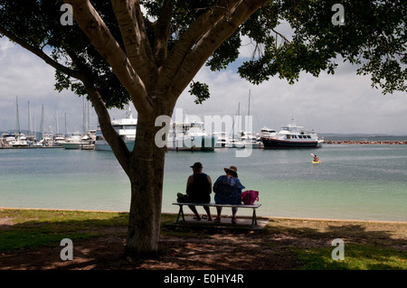 Ein paar sitzen im Schatten eines Baumes mit Blick auf den Yachthafen in Nelson Bay, Port Stephens, New-South.Wales, Australien. Stockfoto