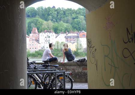 Ein junges Paar sitzt auf einer niedrigen Mauer blickt in den Inn, Passau, Deutschland. Stockfoto