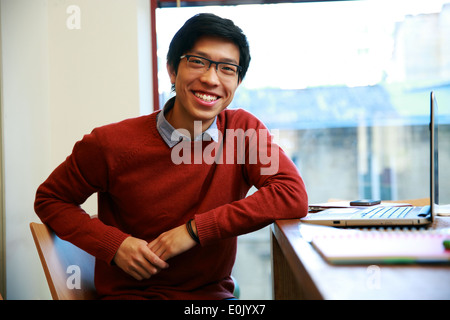Glücklich asiatischer Mann mit Laptop am Tisch sitzen und Blick in die Kamera Stockfoto
