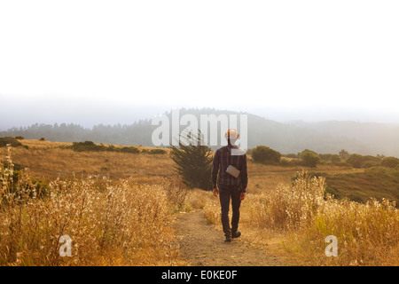 Ein Mann mit einem Strohhut und einem Land Kamera geht auf einen Weg in einem Feld von golden Trockenrasen. Stockfoto