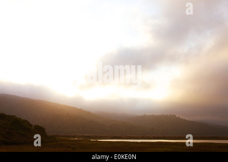 Die Sonne geht über dem nebligen Landschaft mit Bergen in der Ferne und ein Körper des Wassers im Vordergrund. Stockfoto