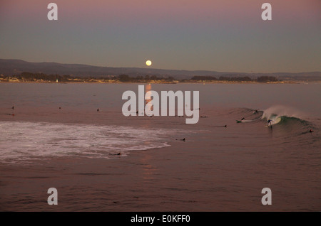 Der Vollmond steigt über dem Pazifischen Ozean am Dampfer, Santa Cruz, während Surfer eine Welle im Vordergrund fangen. Stockfoto