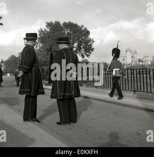 1950er Jahre, historisch, zwei Yeoman Warders, zeremonielle Wächter des Tower of London, die zusammen stehen, London, England, Großbritannien, allgemein bekannt als Beefeaters. Sie gehen auf das Jahr 1485 zurück, als sie von König Heinrich VII., dem Monarchen der Tudor, gebildet wurden. Ein Wachmann der Königin mit Bärenfellhut marschiert im Hintergrund. Stockfoto