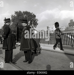 1950er Jahre, historisch, zwei Yeoman Warders, zeremonielle Wächter des Tower of London, die zusammen stehen, London, England, Großbritannien, allgemein bekannt als Beefeaters. Sie gehen auf das Jahr 1485 zurück, als sie von König Heinrich VII., dem Monarchen der Tudor, gebildet wurden. Ein Wachmann der Königin mit Bärenfellhut marschiert im Hintergrund. Stockfoto