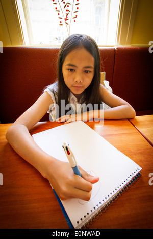 Junge Englisch / Thai Mischlinge Mädchen zeichnen mit einem Stift in einem Malbuch an einem Tisch in einem restaurant Stockfoto