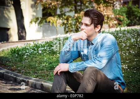 Junger Mann sitzt im Freien im öffentlichen Park, mit der Hand auf seinem Kinn Weg zu denken Stockfoto