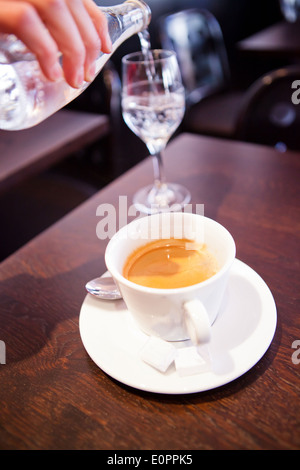 Nahaufnahme der Tasse Espresso-Kaffeemaschine auf hölzernen Café-Tisch und gießt Wasser in Glas Flasche im Hintergrund Stockfoto