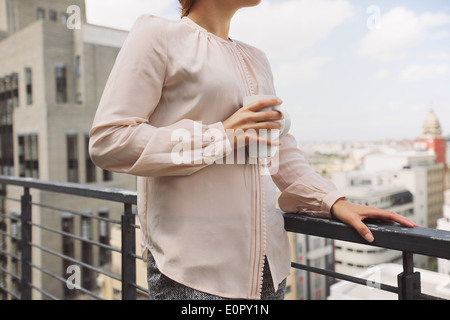Bild der Frau, die ein Kaffee Becher steht auf einem Balkon mit Blick auf Stadt abgeschnitten. Stockfoto