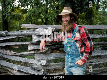 Ein bärtiger Berg-Mann im karierten Hemd und Jeans Overall entspannt durch Split Zaun in eine lebendige Geschichte Staatspark in den Ozarks Arkansas, USA. Stockfoto