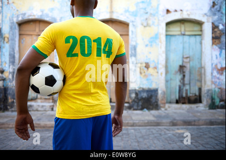 Brasilianischer Fußballspieler in 2014 Shirt steht hält einen Fußball auf einem alten Favela in ländlichen Dorf Straße Stockfoto
