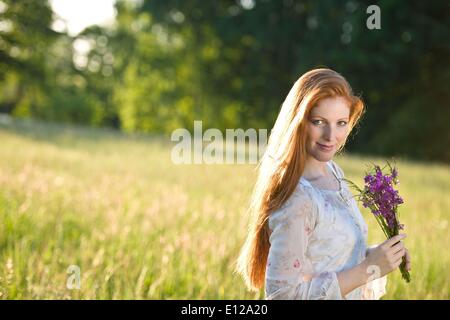 5. Juni 2010 - 5. Juni 2010 - lange rote Haare Frau im romantischen Sonnenuntergang Wiese mit Blumenstrauß Stockfoto