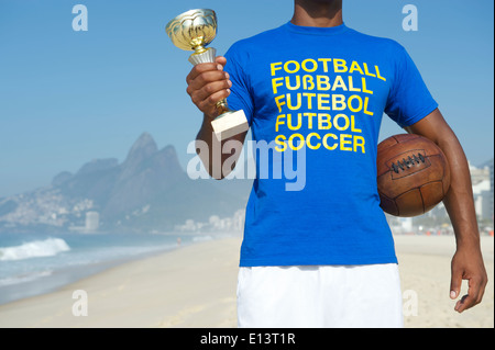 Champion brasilianischer Fußballspieler holding Trophy und Vintage Fußball im internationalen Fußball T-shirt Ipanema Strand Rio Stockfoto