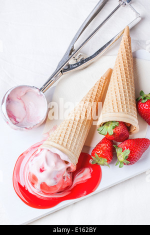 Wafer-Kegel mit Erdbeer Eis mit Sirup, Minze und frischen Erdbeeren serviert auf weiße Schneidebrett auf weißen Textilien. Stockfoto