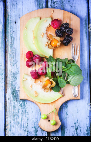 Mischung aus Himbeeren, Brombeeren, Minze, Physalis und Melone auf Holzbrett über lila Holztisch. Ansicht von oben. Stockfoto