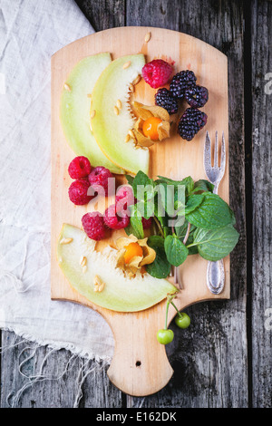 Mischung aus Himbeeren, Brombeeren, Minze, Physalis und Melone auf Holzbrett über alten Holztisch. Ansicht von oben. Stockfoto