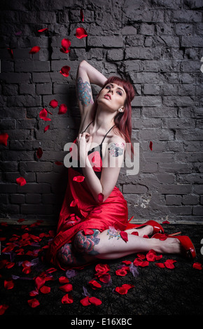 Tätowierte Frau in einem roten Kleid mit roten Schuhen setzt durch eine Mauer, während Rosenblüten auf sie fallen. Stockfoto