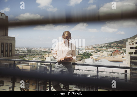 Bild durch Fenster Jalousien von einer jungen Frau, die mit digital-Tablette auf einen Balkon mit Blick auf Stadt Stockfoto