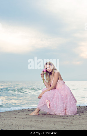 Eine schöne junge Frau Porträt sitzen am Strand, gekleidet in einem formalen rosa 50er Jahre Kleid, hält eine Rose auf ihrem Gesicht. Stockfoto