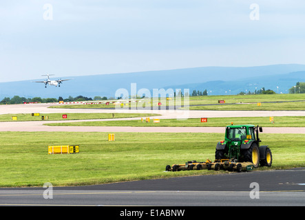 Traktor Pulling Rasenmäher Ausschnittgras am Manchester International Airport mit Flybe Q400 Flugzeug Landung in England Hintergrund Stockfoto