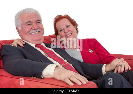 Kaukasische älteres paar Lächeln auf den Lippen und Hand in Hand, tragen elegante Kleidung, sitzend auf einer bequemen roten couch Stockfoto