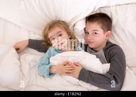 Liebevolle kleine Bruder und Schwester im Bett zusammen Kuscheln unter der Bettdecke bis verschlafen in die Kamera schaut Stockfoto