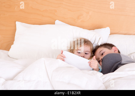 Kleinen Bruder und Schwester Kuscheln bis zusammen vor dem Schlafengehen unter eine warme Bettdecke, wie sie ein e-Book lesen oder Surfen Sie im Internet auf eine t Stockfoto